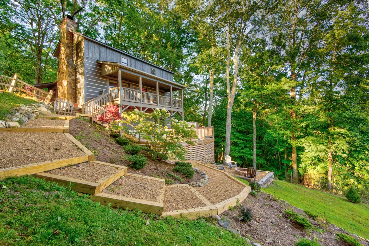A welcoming view of a wooden house set amid terraced gardens, with a deck featuring a railing and surrounded by mature trees. The fully fenced yard is visible, providing open, secure space perfect for pets to play outdoors. The tranquil environment suggests a safe and enjoyable place for families and their dogs to relax together.