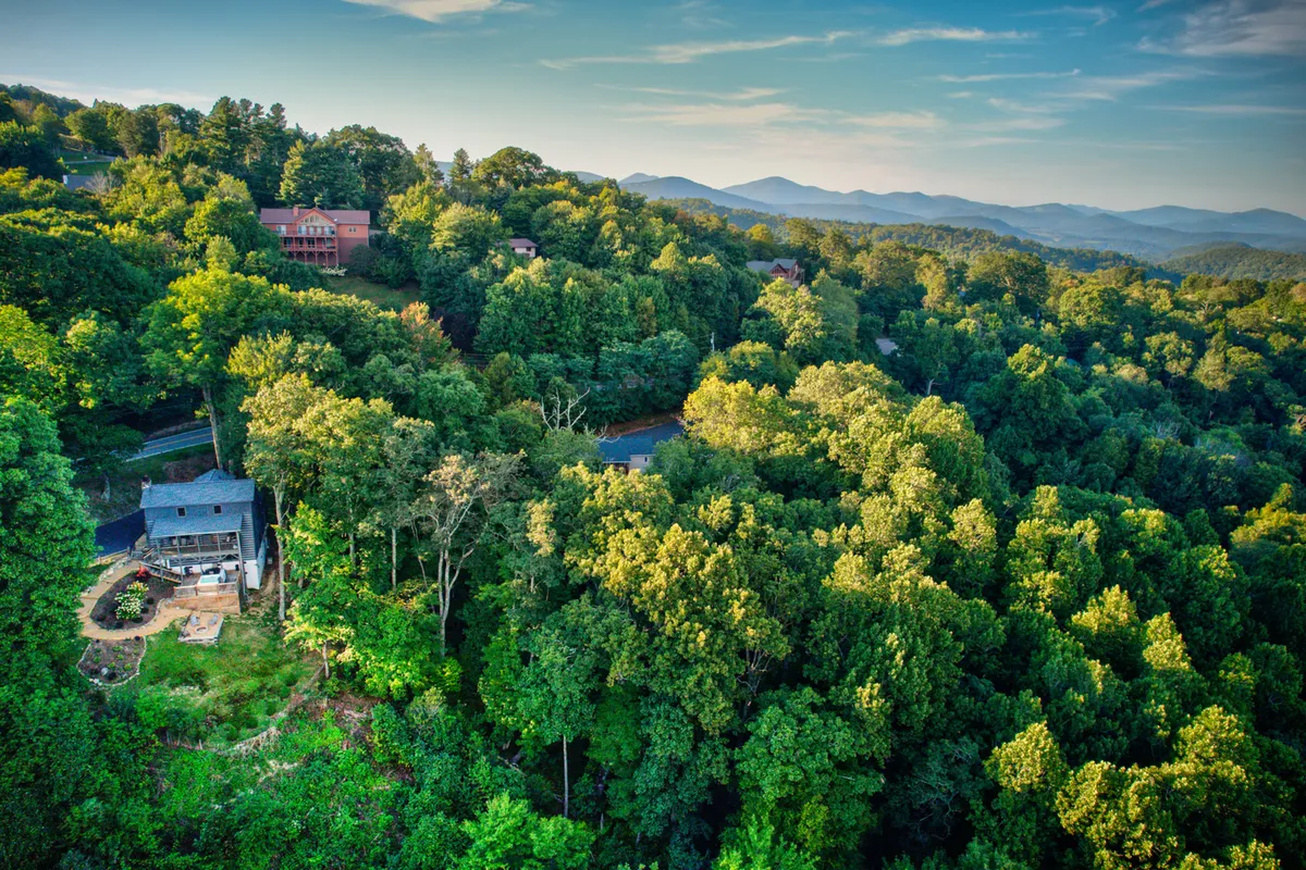 A peaceful exterior view featuring lush greenery, a mountainous backdrop, and a few homes nestled in the trees. The patio area is visible, inviting relaxation and social gatherings in a natural setting. The landscape evokes the quiet beauty of mountain hiking destinations.