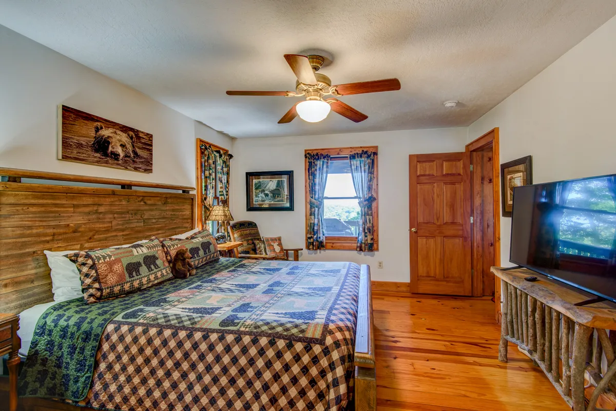A welcoming bedroom with a wooden bed frame, a colorful quilt, matching wooden furniture, and a window with curtains. Soft light fills the room, and framed artwork adds a personal touch, making the space inviting and warm.