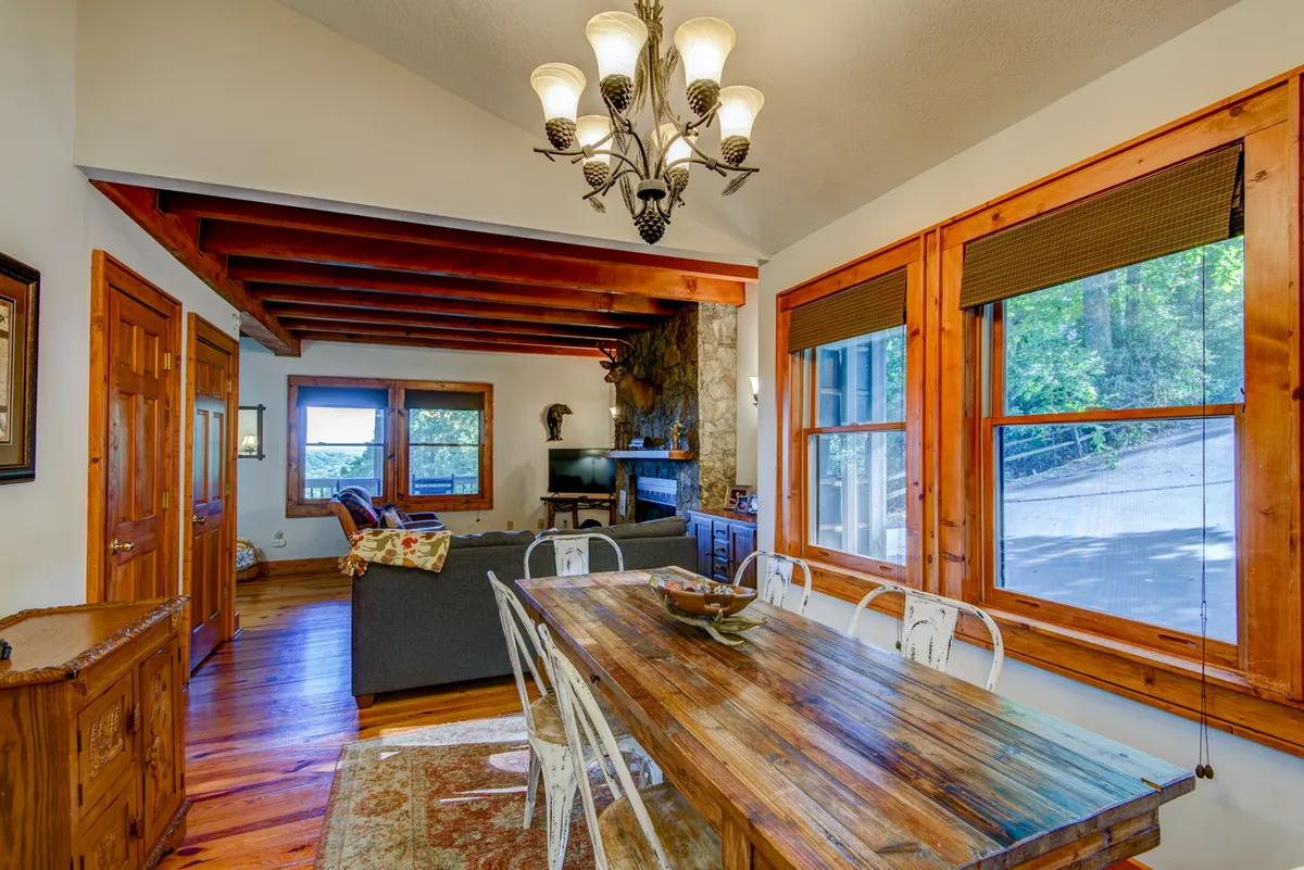 A cozy dining area featuring a substantial wooden dining table, an elegant chandelier above, large windows letting in natural light, and a prominent stone fireplace as a backdrop. Plush seating around the table invites guests to linger and enjoy meals or games beside the warmth of the fire.