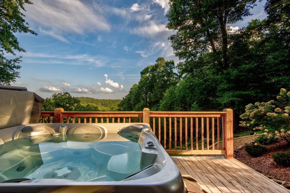 This image showcases a private hot tub on a wooden deck surrounded by lush greenery and distant hills, with ample space for relaxation and unobstructed views of the natural landscape. The tranquil mood and scenic setting highlight the ideal environment for couples seeking privacy and romance.