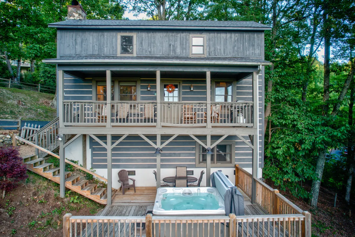 A hot tub sits invitingly on a wooden deck, which also features outdoor seating areas and is surrounded by trees. The image conveys warmth, comfort, and a sense of privacy amidst nature—perfect for unwinding after a day in the snow.