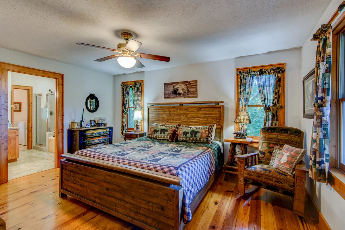 A cozy bedroom with plaid bedding, wooden furniture, decorative bear pillows, and warm lamps on the nightstands. Framed artwork hangs above the bed, and sunlight streams softly through the windows, creating a restful atmosphere.