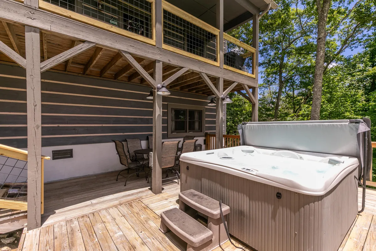 A lower wooden deck with a bubbling hot tub and an alfresco dining set, surrounded by lush trees. This space invites relaxation with the hot tub and is perfect for meals outdoors, echoing the narrative's focus on unwinding after adventures and enjoying the fresh mountain air.