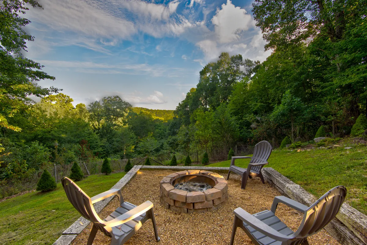 Adirondack chairs are arranged around a fire pit in a wooded outdoor setting. The scene is cozy and inviting, perfect for gathering, storytelling, or warming up after a day outdoors.