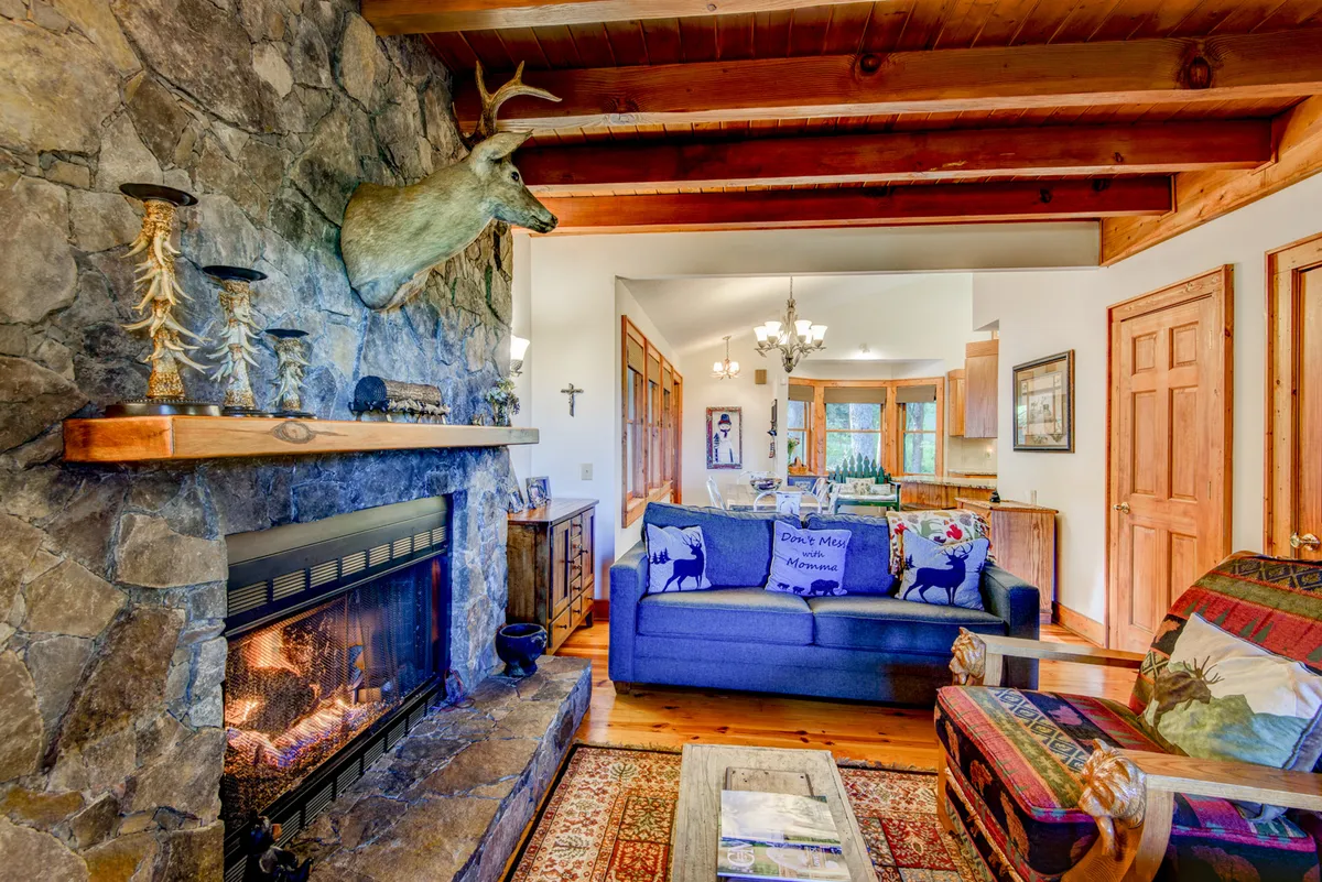 A mountain-style living room featuring a rustic stone fireplace, blue sofa, wooden beams, and a colorful armchair. The space is arranged for comfort and sociability, with a mounted deer head and cozy decor enhancing the inviting, lodge-like atmosphere.
