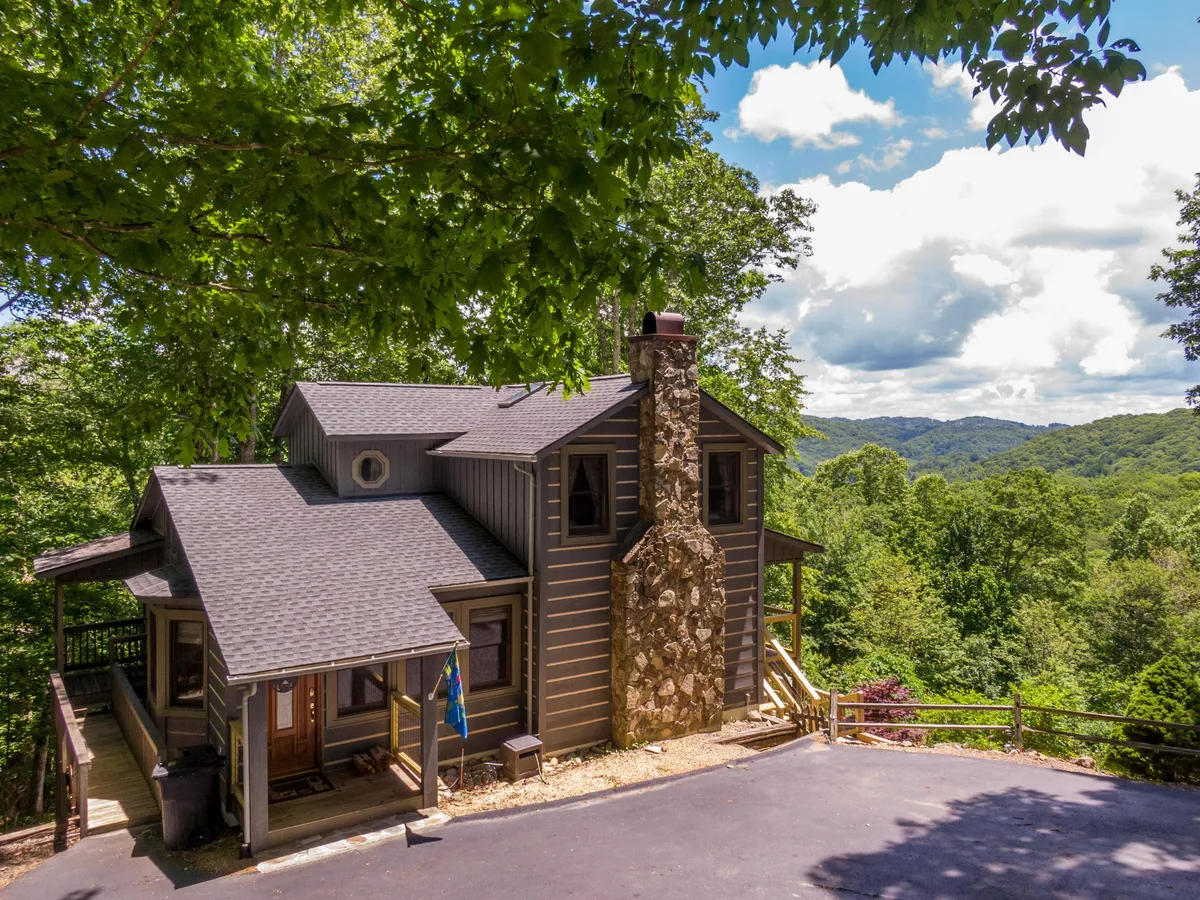 This image features a wooden deck attached to the home, a prominent stone chimney, surrounding trees, and sweeping mountain views in the background. The scene feels peaceful and showcases the outdoor living experience close to nature.